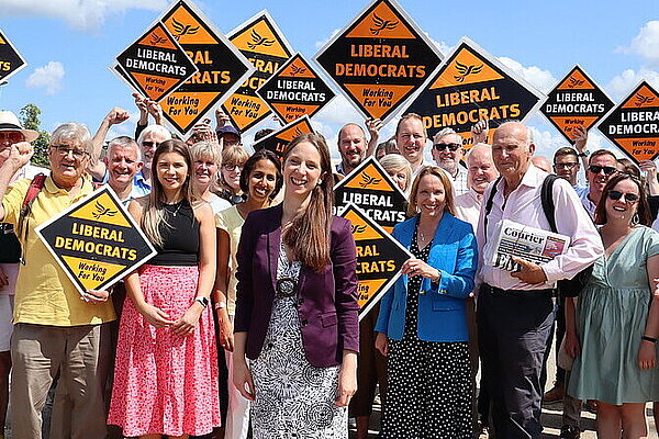 A group of Liberal Democrats holding gold diamond Liberal Democrat posters behind Mid Bedfordshire candidate Emma Holland-Lindsey